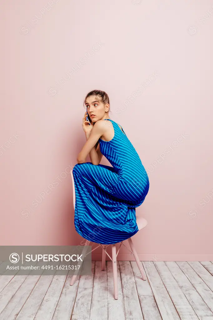 Side view of young woman crouched on top of a chair looking away while talking on the mobile phone on pink background