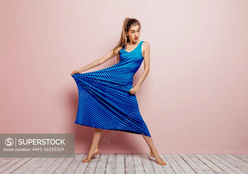 Young woman barefoot looking away stretching side of a blue dress while standing on minimalist pink background