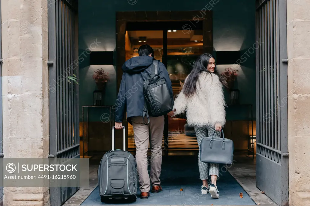 Back view of happy stylish young couple in love with suitcase and bags holding hands and entering modern city hotel during travel while woman looking at camera over shoulder
