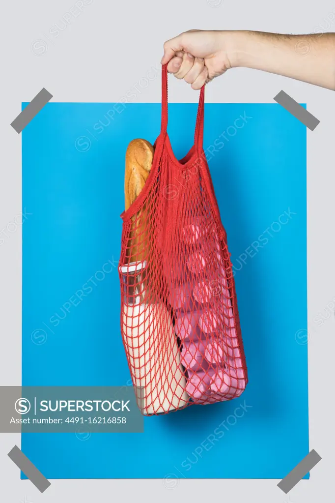 Unrecognizable person carrying net bag with various groceries against blue rectangle during zero waste shopping