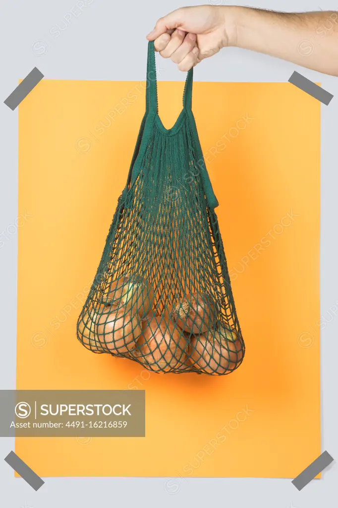 Anonymous person carrying green string bag with ripe onions against yellow rectangle during eco friendly shopping
