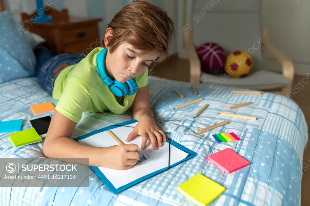 Thoughtful schoolboy in casual wear and wireless headphones enjoying music and drawing with pencils while spending free time in bedroom