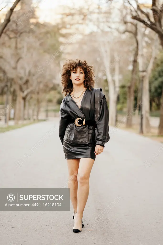 Optimistic female entrepreneur in trendy leather jacket and with curly hair smiling at camera while standing on blurred background of street