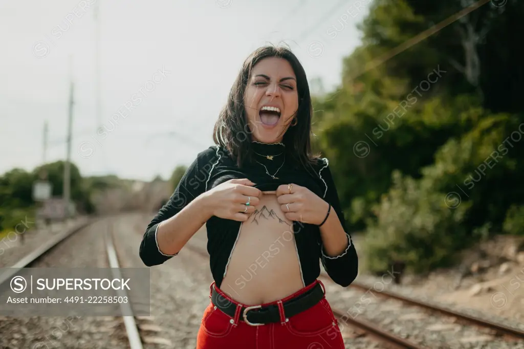 Pretty young woman in stylish outfit screaming and lifting sweater to show mountains tattoo while standing on blurred background of rails