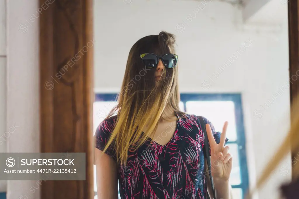 Pretty woman standing at mirror with sunglasses over hair and gesturing two fingers.