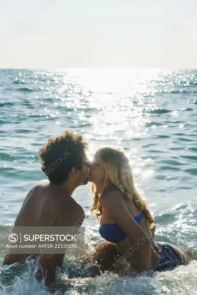 Loving couple swimming in blue water of ocean and kissing in bright back light.