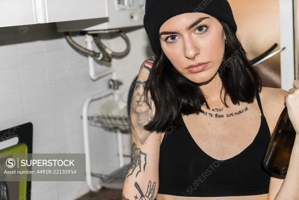 Young tattooed brunette in hat and black bra holding glass bottle and looking at camera on kitchen.