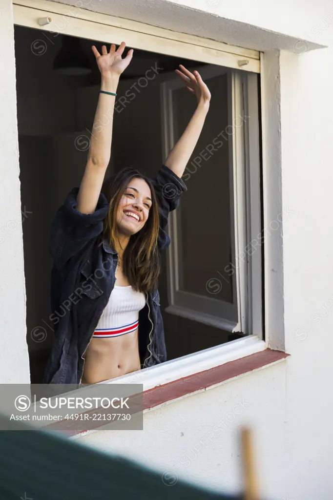 Young beautiful girl in sportive bra and denim shirt smiling and stretching in open window.