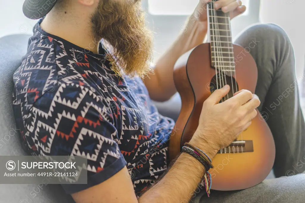 Bearded man sitting in armchair and playing guitar at home.