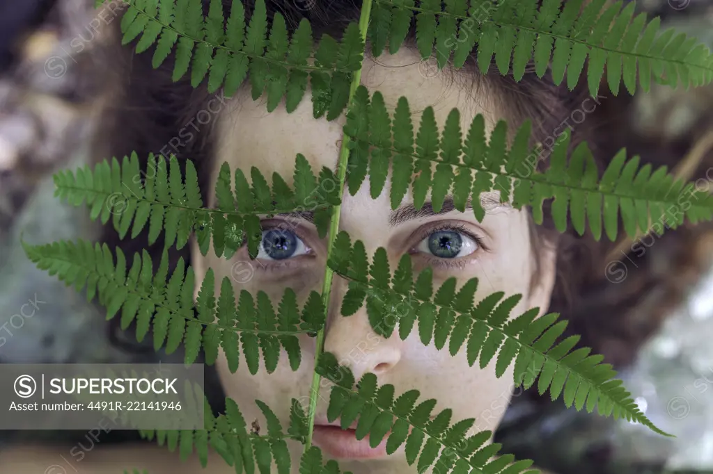 From above shot of girl looking at camera while covering face with green fern leaf.