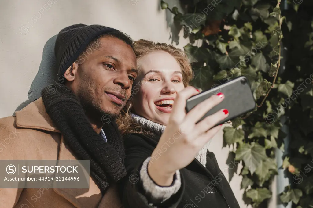 Happy multiethnic couple in warm clothes standing at bush and taking selfie.