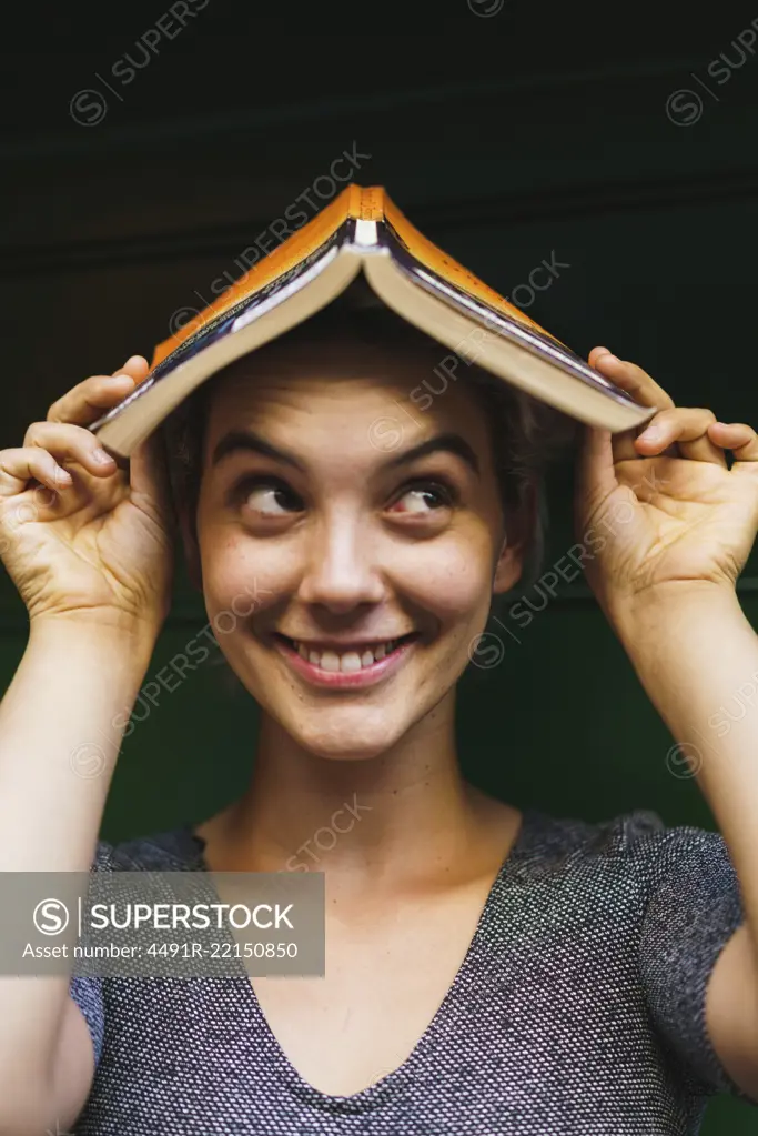 Portrait of young playful woman smiling away and holding opened book on head.