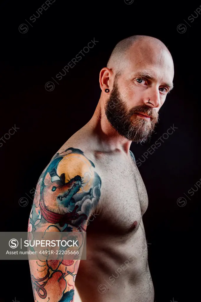 Side view of shirtless bald serious hipster with tattoos on hands looking at camera on black background