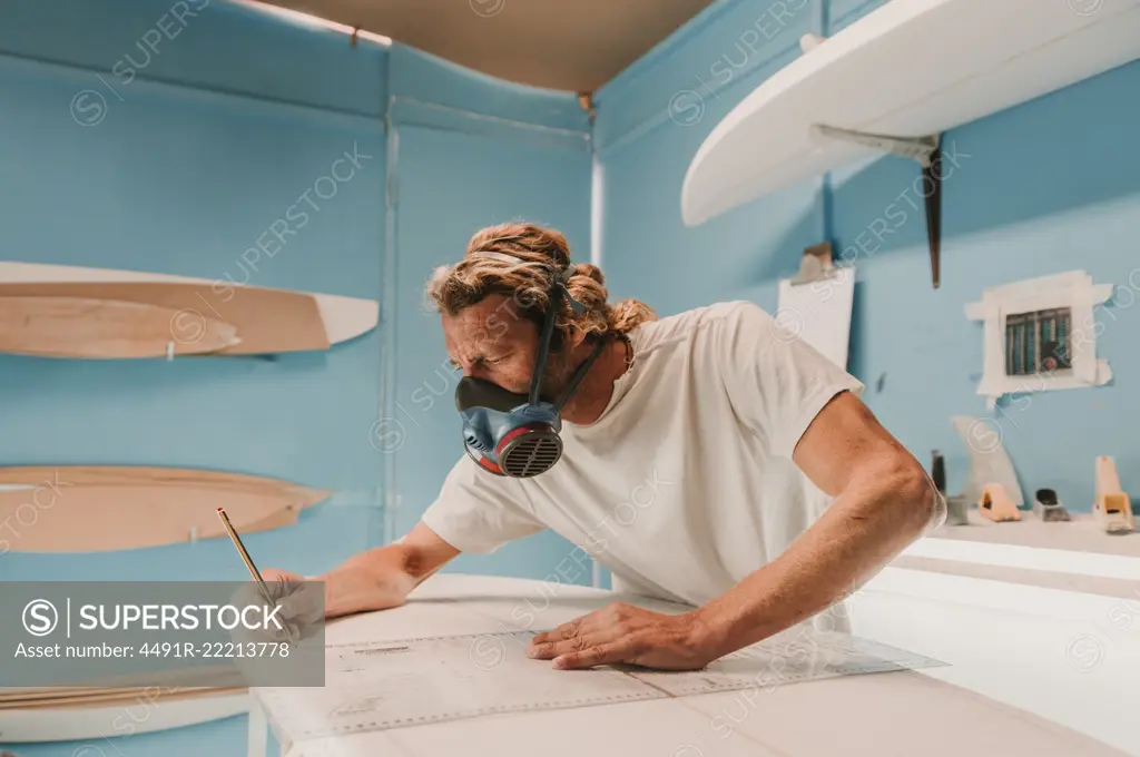 Side view of male in breather holding pencil and measuring surf board in workplace