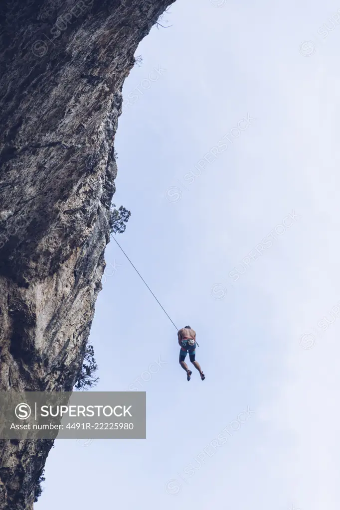 From below unrecognizable climber hanging on rope on rough cliff against blue sky