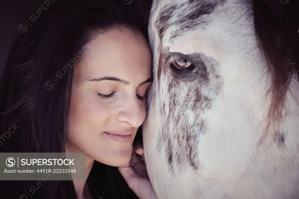 Attractive charming woman with closed eyes touching beautiful horse in stall