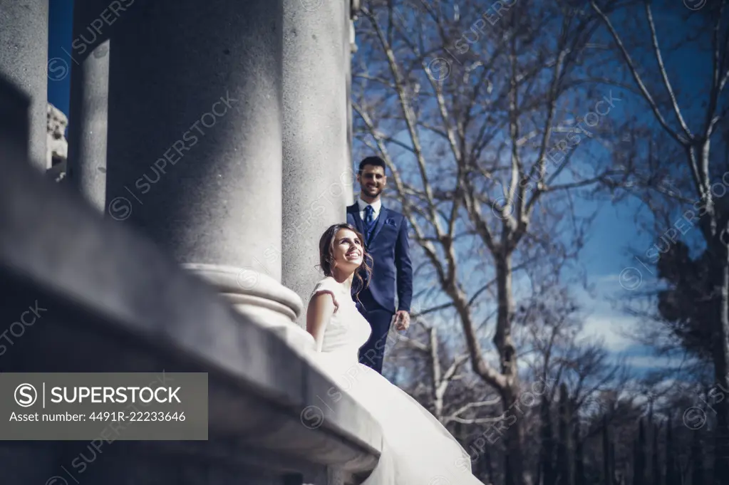 From below side view of young elegant couple in wedding dresses near columns on street in sunny day