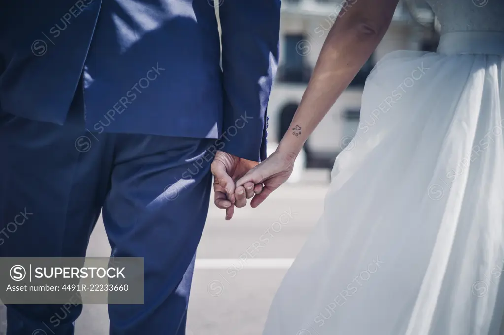 crop man holding on hands woman in wedding dress on street in sunny day