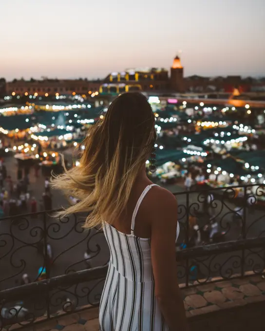Back view of trendy woman with flying hair standing on balcony against bright lights of Moroccan city in twilight