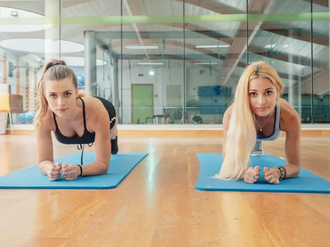 Two muscular slim women in sportswear looking at camera while doing plank on mat in gym