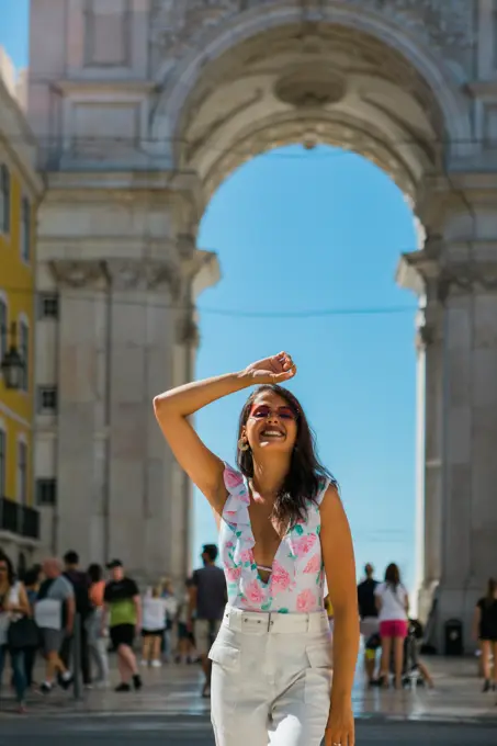 Young happy woman in sunglasses standing beside majestic arch in city street in Lisbon, Portugal