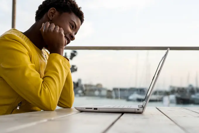Black African American woman in yellow jacket using laptop at wooden desk in city on blurred background 
