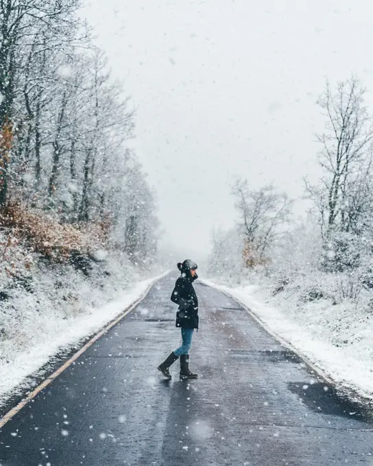 Side view of lonely female in black jacket and jeans crossing empty country road on snow and gloomy winter day