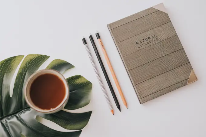 From above stylish notebook with wooden cover composed with row of pencils and mug of hot drink on green Monstera leaf on white background