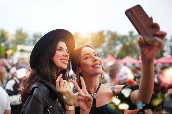 Charming cheerful friends in black hat having fun grimacing and taking selfie on mobile phone in bright day at festival