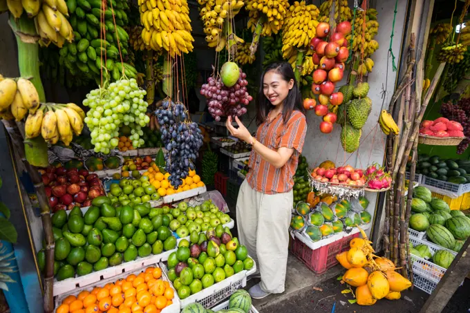 From above of Asian pleased woman on vacation in casual clothing making choice of fresh fruits at market at Sri Lanka;Satisfied Asian resting lady in casual wear tasting grape and smiling at outdoors market