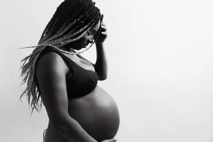 Cheerful young pregnant black female with braids touching belly and looking away with smile while standing against white wall