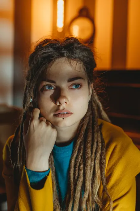 Stylish millennial female with dreadlocks wearing yellow coat over blue sweater looking away thoughtfully while sitting in room with piano in background;Pensive hipster woman looking away
