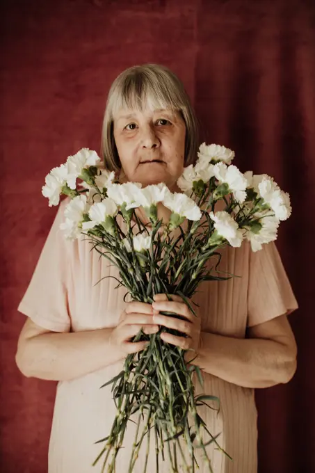 From above thoughtful old female with gray hair looking at camera and holding bouquet of white carnation at home