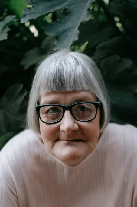 Senior woman with grey hair looking at camera in eyeglasses among plants with big green leafs