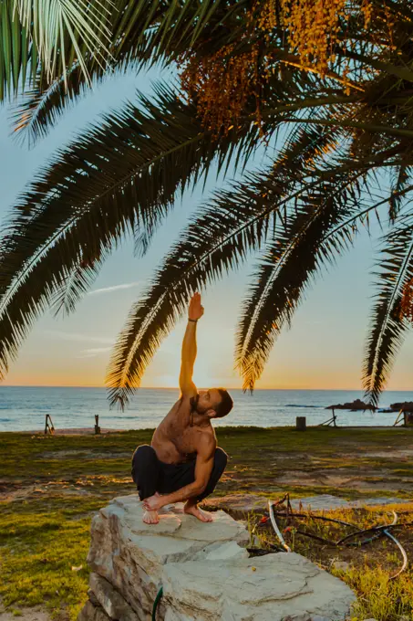 Shirtless man athlete doing yoga poses on rocky fence with empty beach blue sea and palm leafs on background during sunset