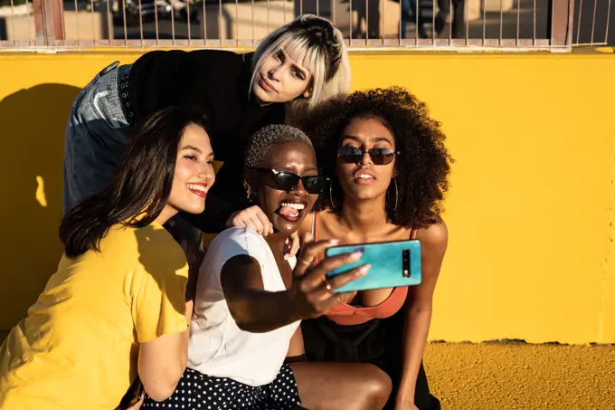 Cheerful young diverse female friends taking selfie on smartphone in street