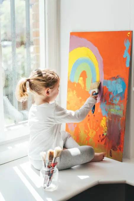 Creative blond girl in casual clothes sitting on window sill against window and painting with paintbrush large multi colored rainbow on orange canvas
