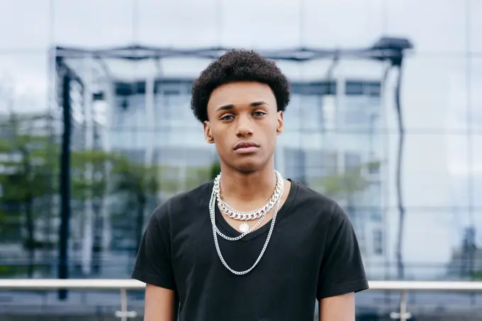 From below of young curly haired African American teenage guy in black t shirt and trendy neck chains looking at camera while standing against blurred modern building with glass wall
