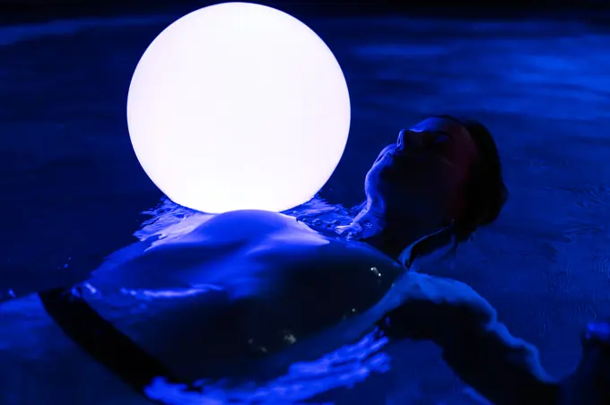 From above of relaxed slim female lying on water in dark swimming pool with luminous ball