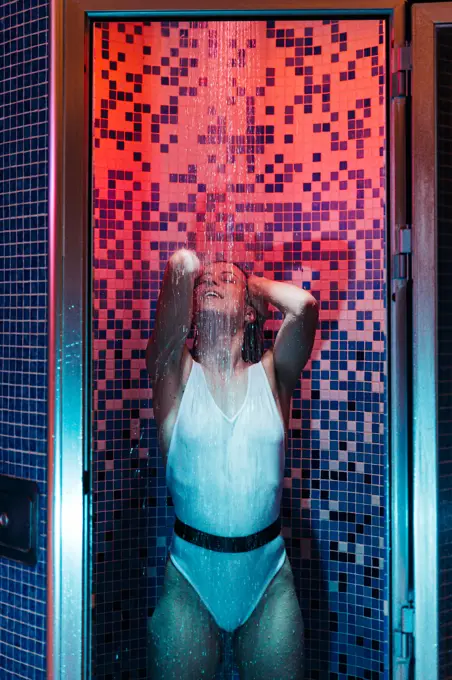 young female in swimwear enjoying water and relaxing in shower cabin illuminated with red and blue light