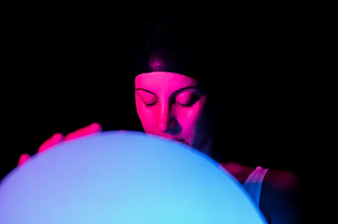 Tranquil young female in swimsuit and cap holding luminous ball while standing in dark swimming pool with neon light