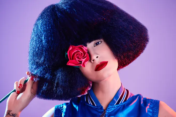 Crop female model in synthetic wig and with red lipstick in a purple background holding a rose