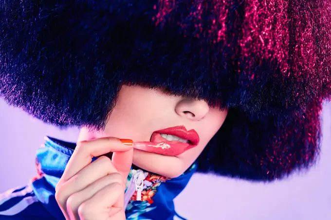 Trendy female in lavish wig removing glossy sticker from lip under magenta light against violet background