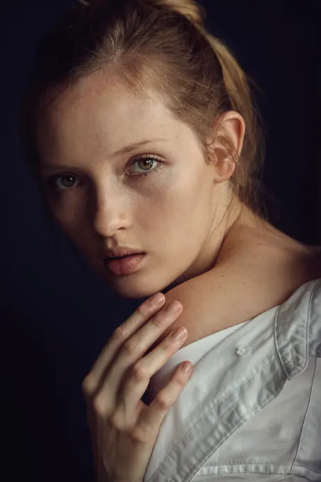 Young lady in white shirt touching delicate skin of shoulder and looking at camera on black background