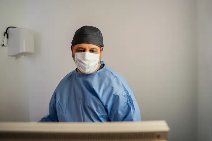 Serious adult doctor in protective masks and medical uniform focusing on screen and analyzing information while working in light operating room of modern hospital