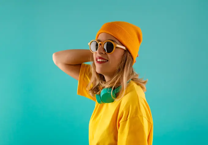 Side view of happy young cute female in yellow outfit and orange beanie looking away wearing sunglasses sunglasses against colorful turquoise background
