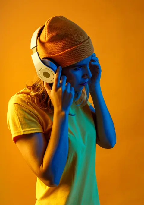 Side view of confident thoughtful young woman in orange beanie listening to music on headphones looking away against orange background