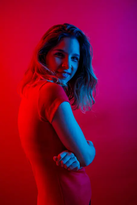Side view of happy young woman with crossed arms smiling and looking at camera while standing under blue neon illumination against red background