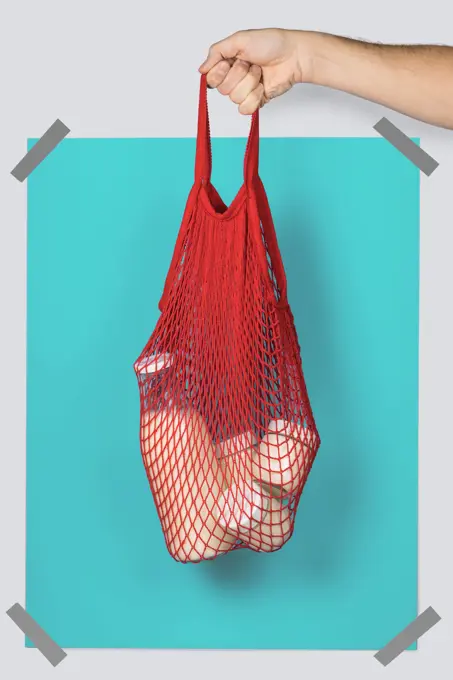 Anonymous person carrying red string sack with glass containers of dairy against turquoise rectangle during eco friendly shopping
