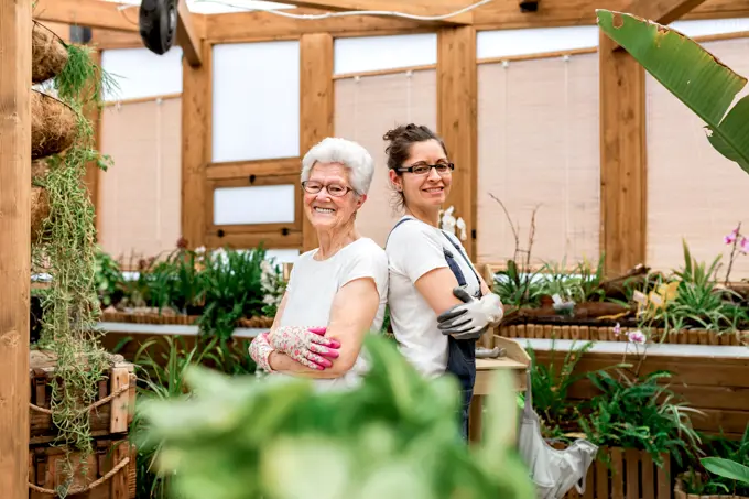 Side view of positive elderly and adult women in gloves and glasses smiling for camera and crossing arms while working in indoor garden together
